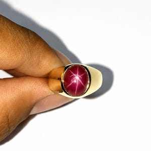 May include: A gold ring featuring a polished, oval-shaped cabochon star ruby gemstone. The deep magenta-red stone displays a distinct six-rayed star effect centered on its surface, set within a smooth, solid yellow gold band with a wide shank.