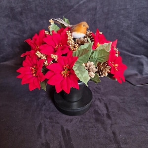 May include: A decorative floral arrangement featuring vibrant red poinsettia flowers, green leaves, small gold-colored berries, and brown pinecones. A small artificial robin bird with an orange breast sits atop the arrangement in a black pot.