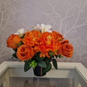May include: A vibrant floral arrangement featuring orange roses and chrysanthemums, accented with white lilies and green foliage. The flowers are arranged in a black pot, creating a striking contrast. This decorative piece adds a touch of color and elegance to any space.
