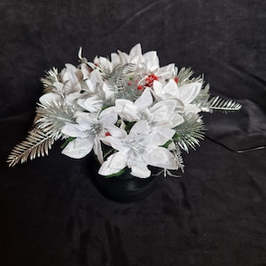 May include: A floral arrangement featuring white poinsettia flowers, silver-toned fern-like foliage, and small red berries. The bouquet is contained within a simple black circular base, creating a high-contrast display of white, silver, and red elements.