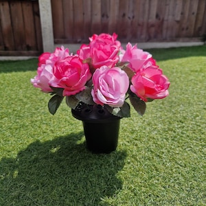 Könnte beinhalten: Ein Strauß aus rosa und fuchsiafarbenen Rosen in einer schwarzen Plastikvase. Die künstlichen Blumen haben grüne Blätter und sind kreisförmig angeordnet. Die Vase steht auf einer grünen Wiese.