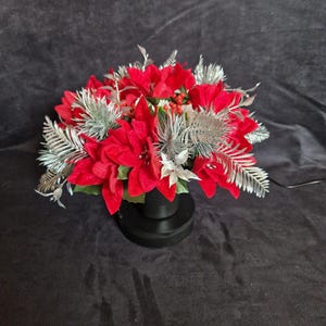 May include: A floral arrangement featuring vibrant red poinsettia flowers interspersed with metallic silver fern-like foliage and small red berries. The arrangement is contained within a simple black cylindrical base, creating a contrast.