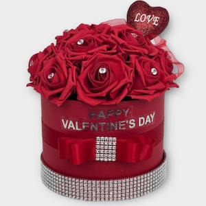 May include: A red round box with a bouquet of red roses, each with a rhinestone center. The box has a red ribbon and bow with a rhinestone center, and a sparkling trim. The text "HAPPY VALENTINE'S DAY" is displayed on the box. A glittery heart with the word "LOVE" is on top.