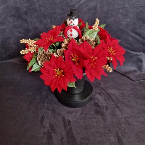 May include: A holiday floral arrangement in a black pot featuring bright red poinsettia flowers, gold-painted pinecones, and gold berry sprigs. A small snowman figurine wearing a black top hat and a red scarf is nestled in the center of the bouquet.