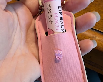 May include: A pink lip balm tube with "Peach" text is held in a pink case with a small, clear, pink gummy bear charm. The lip balm is labeled "Lip Balm, Moisturizing & Soothing, Tamper Sealed". The case has a silver key ring.