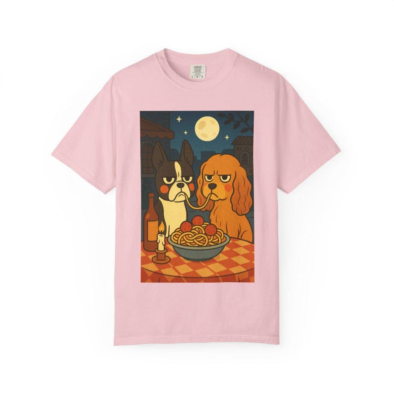 Puede incluir: Camiseta rosa claro con una ilustraci&oacute;n de dibujos animados de dos perros compartiendo espaguetis. La escena incluye una botella de vino, una vela encendida y una luna llena en el cielo nocturno. Los perros tienen expresiones de mal humor.