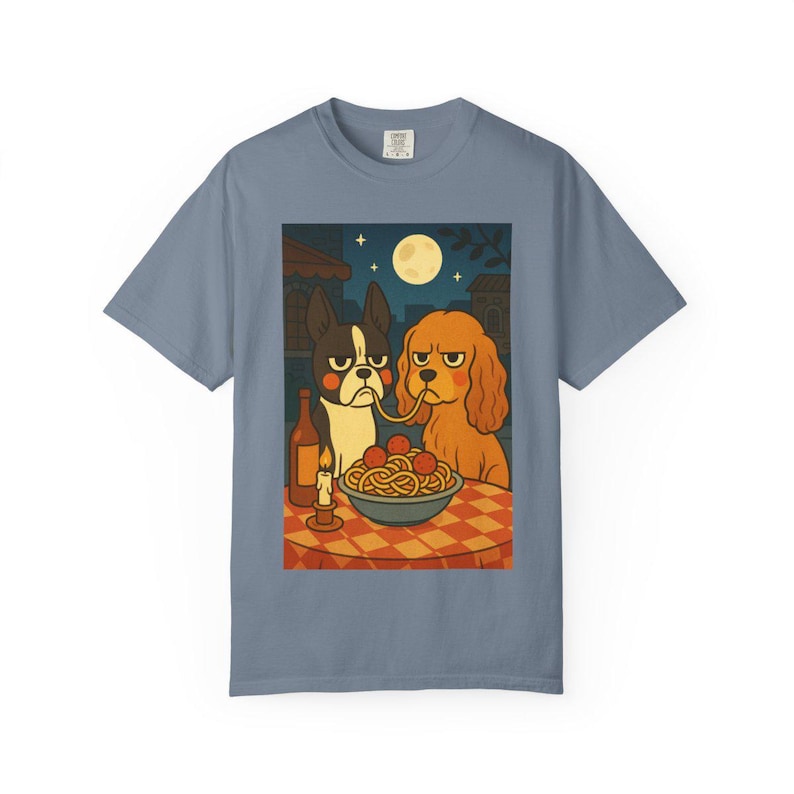 Puede incluir: Camiseta azul con una ilustraci&oacute;n de dibujos animados de dos perros compartiendo espaguetis. La escena incluye una botella de vino, una vela encendida y una luna llena en el cielo nocturno. Los perros tienen expresiones gru&ntilde;onas.