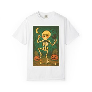 May include: White t-shirt featuring a Halloween-themed graphic. The design depicts a dancing skeleton in a graveyard with pumpkins, candles, and a crescent moon. The skeleton is holding a top hat.
