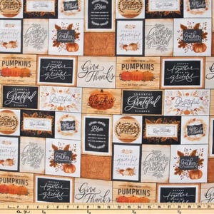 May include: A cotton fabric featuring a repeating pattern of rectangular and circular blocks with autumn themes, including orange pumpkins, fall foliage, and text such as Gather, Grateful, and Give Thanks, with a yellow measuring tape at the bottom.