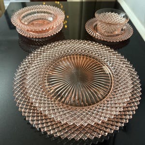 May include: Pink glass dinnerware set with a diamond pattern. The set includes plates, bowls, and teacups with saucers. The dishes are arranged on a black surface, showcasing their intricate design and color.