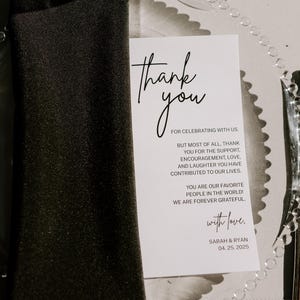 Könnte beinhalten: Eine Hochzeitstafel mit einer schwarzen Serviette, einem durchsichtigen, mit Perlen besetzten Platzteller und einer Dankeskarte. Die Karte lautet "Danke" und enthält eine Nachricht sowie die Namen Sarah & Ryan mit dem Datum 04.25.2025. Ein Weinglas und Besteck sind ebenfalls vorhanden.