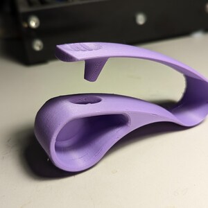 Peut inclure: Objet incurvé imprimé en 3D, de couleur lavande, avec un design unique. L'objet a une texture lisse et un trou au centre. Le design comprend une section surélevée qui se courbe vers le haut. Il s'agit probablement d'un outil ou d'un objet décoratif.