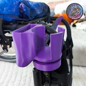May include: A bright purple cup holder, designed for attachment to a wheelchair or mobility aid. The holder has a circular base and a dual-compartment design, with a smooth, glossy finish. It is mounted on a black frame.