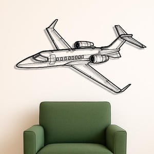 May include: A black metal wall art piece depicting a detailed outline of a private jet. The artwork is mounted on a white wall above a green armchair with wooden legs. A black pillow rests on the wooden floor.