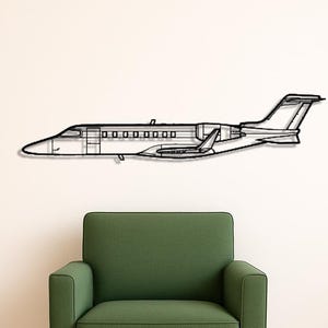 May include: A black metal wall art piece depicting a detailed outline of a private jet. The artwork is mounted on a white wall above a green armchair with wooden legs. A black pillow rests on the wooden floor.