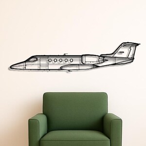 May include: A black metal airplane wall art piece is mounted on a white wall above a green armchair. The airplane is a detailed outline of a jet, with visible windows and engine details. The chair has wooden legs and a black pillow.