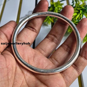 May include: A smooth, circular silver bangle bracelet with a polished, reflective metallic finish. The bangle features a simple, continuous band design without any additional embellishments or patterns. The text saijewelleryhub is visible.