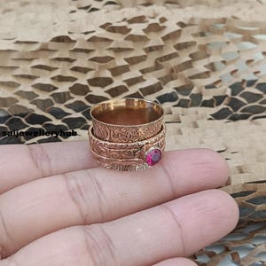 May include: A gold-colored spinner ring with intricate floral and scrollwork designs. The ring features a central band that spins, and a small, round, red gemstone is set on the side. The ring is displayed on a hand.