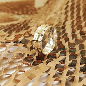 May include: A silver ring with a gold band, set against a textured brown background. The ring has a polished finish and a simple, elegant design. The ring is a piece of jewelry.