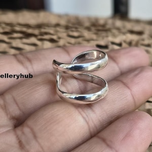 Könnte beinhalten: Ein silberfarbener Ring mit einem einzigartigen Doppelband-Design. Der Ring wird in einer Hand gehalten und zeigt seine polierte Oberfläche und geschwungene Form. Das Design des Rings ist modern und auffällig, geeignet für den Alltag. Der Text "Saijewelleryhub" ist sichtbar.