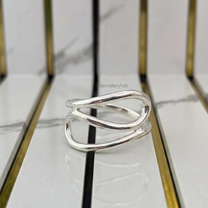 Può includere: Un anello in argento lucido caratterizzato da un design fluido e traforato con anelli sovrapposti. La superficie metallica è liscia e riflettente, creando un aspetto stratificato. Il testo 'saijewelleryhub' è visibile sullo sfondo.