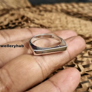 Puede incluir: Un anillo de plata con una cara rectangular. El anillo se muestra en la mano de una persona. El texto "Saijewelleryhub" es visible en la imagen. El anillo es de diseño sencillo y moderno.