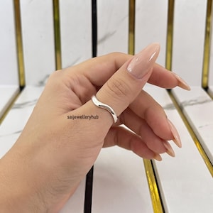 May include: A polished silver ring with a smooth, undulating wave-like design worn on the thumb. The metal has a reflective surface and a continuous, flowing shape that wraps around the finger. The text saijewelleryhub is visible.