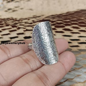 Op de afbeelding: Een zilverkleurige ring met een langwerpig, schildachtig ontwerp. De ring heeft een ingewikkeld, in reliëf aangebracht bloemmotief over het oppervlak. De ring wordt op een hand getoond, met de merknaam "Saijewelleryhub" zichtbaar.