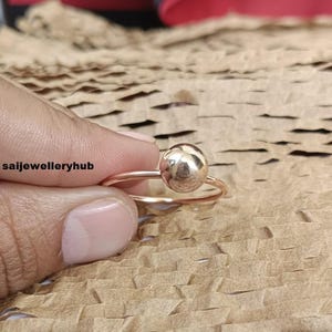 May include: A rose gold ring with a polished sphere as its central feature. The ring has a thin band, and the sphere is the main focus. The ring is set against a textured, brown paper background. The text "saijewelleryhub" is visible.