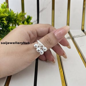 May include: A silver ring featuring a band adorned with multiple small, dangling spherical charms. The ring is displayed with the text saijewelleryhub visible in the background. The metallic surface of the ring and the attached round beads reflect light, creating a polished and bright appearance on the finger.