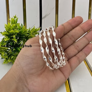 Könnte beinhalten: Ein silberfarbenes Spiralarmband mit einem durchgehenden, gedrehten und gewellten Design, das sich mehrfach umwickelt. Die metallische Oberfläche ist poliert und reflektierend und bildet eine Reihe von miteinander verbundenen Schlaufen.
