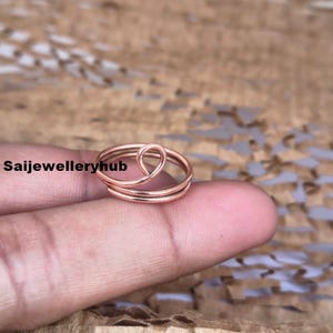 Könnte beinhalten: Ein roségoldener Ring mit einem einzigartigen Design. Der Ring hat ein dreifaches Band mit einem geschlungenen Akzent oben. Der Ring wird auf einem Finger vor einem strukturierten, braunen Hintergrund gezeigt. Der Ring ist ein Schmuckstück.