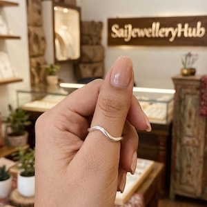May include: A silver-colored ring with a wavy design is shown on a finger. The ring has a smooth, polished finish and a simple, elegant style. The background includes a jewelry store setting with display cases and signage.