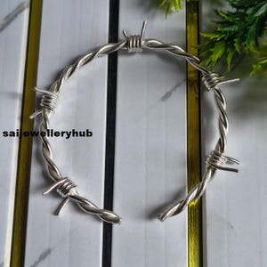 Op de afbeelding: Een zilverkleurige metalen armband ontworpen als een stuk prikkeldraad, met gedraaide metalen segmenten en scherpe, uitstekende punten langs de cirkelvormige band. De tekst saijewelleryhub is zichtbaar aan de linkerkant.