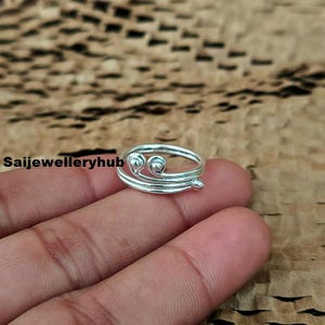 以下が含まれることがあります： スパイラルデザインのシルバーリング。リングには、2つの小さな丸い装飾とコイル状のバンドが付いています。リングは、テクスチャード加工された背景に対して指に表示されています。「Saijewelleryhub」というテキストが表示されます。