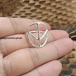 May include: A silver ring with a peace symbol design. The ring has a simple, open design with a circular outer shape and three interior lines forming the peace symbol. The ring is displayed on a hand.