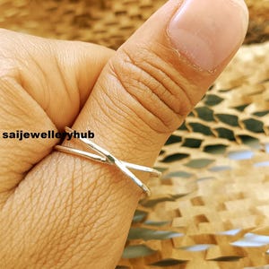 Peut inclure: Une bague argentée avec un design en forme de X est portée au doigt. La bague est simple et élégante, avec une esthétique minimaliste. Le fond est une surface texturée, brun clair. Le texte "saijewelleryhub" est visible en bas à gauche.