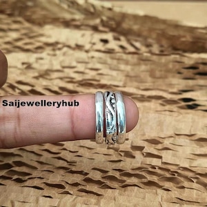 Puede incluir: Tres anillos de plata apilados en un dedo. El anillo central tiene un diseño grabado detallado, flanqueado por dos bandas lisas. Los anillos se muestran sobre un fondo marrón texturizado. El texto "Saijewelleryhub" es visible.
