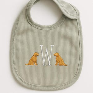 May include: Sage green baby bib with a ribbed texture. The bib features an embroidered capital letter "W" flanked by two golden retriever dog illustrations. The bib has a rounded shape with a curved neck opening.