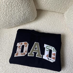 May include: Black sweatshirt with the word "DAD" in a collegiate style. The letter "D" features a football pattern, the "A" is olive green, and the second "D" has a football and the word "are" in white.