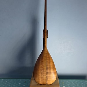 May include: A wooden stand-up paddleboard paddle with a teardrop-shaped blade and a T-shaped handle. The paddle is made of light brown wood and is mounted on a wooden base. The paddle is designed for stand-up paddleboarding.