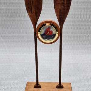 May include: A wooden desk clock featuring a clock face with a landscape design, flanked by two wooden paddles. The clock is set on a rectangular wooden base. The paddles and base have a rich, dark brown color.