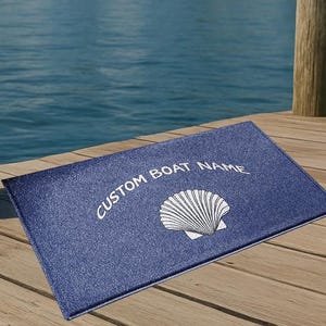 May include: A blue rectangular boat mat with the text "CUSTOM BOAT NAME" and a white seashell design. The mat is on a wooden dock near a body of water with houses in the background.