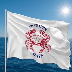 May include: A white flag with a red crab illustration and the words "NO GRABBY DAYS" in blue. The flag is waving in the wind against a blue sky and ocean background.