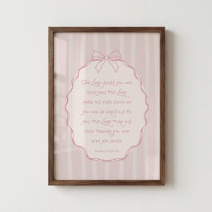 May include: A framed print with a pink and white striped background. The print features a pink oval with a bow at the top, containing the text "The Lord bless you and keep you..." in pink script. The frame is a dark brown color.