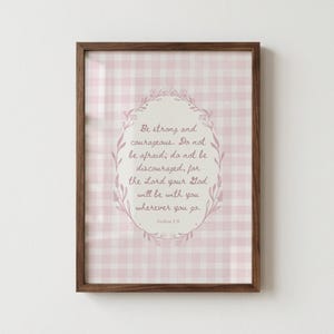 May include: A framed print with a pink and white gingham pattern background. The print features an oval floral design with the text "Be strong and courageous..." in a script font. The frame is a dark brown colour.