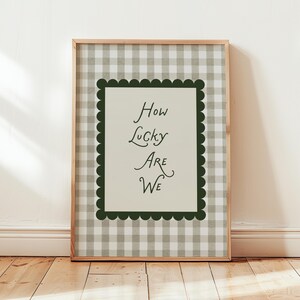 Könnte beinhalten: Ein gerahmter Druck mit dem Text "How Lucky Are We". Der Druck hat einen hellgrünen und weißen Vichy-Karo-Hintergrund. Der Text ist in einer Schreibschrift gehalten und der Druck ist mit einem wellenförmigen dunkelgrünen Rand und einem hellbraunen Holzrahmen gerahmt.