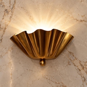 May include: A semi-circular wall sconce with a metallic gold finish and a pleated, fan-like design. The fixture features a small spherical accent at the base and emits a warm, upward-directed light that creates a soft glow against the wall surface.