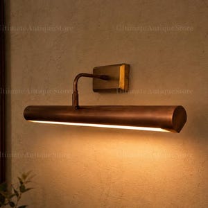 May include: A wall-mounted picture light with a cylindrical, metallic copper-toned shade and a rectangular brass-coloured base. The fixture features a curved arm connecting the shade to the wall plate, emitting a warm, downward-facing light.