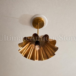 May include: A gold-colored, ruffled ceiling light fixture with a single exposed bulb. The fixture is suspended from a brass rod and a white ceiling plate. The light has a unique, decorative design.